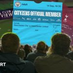 A group of fans are sitting in the stands at a football watch, in front of them are a number of large scale tickets for Manchester City, Everton, West Ham, and Arsenal
