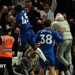 Enzo Maresca celebrating with a huddle of Chelsea players after the winner against Liverpool, as Romeo Lavia leaps into the air and punches the air towards the crowd of supporters, who are all smiling and celebrating in the background